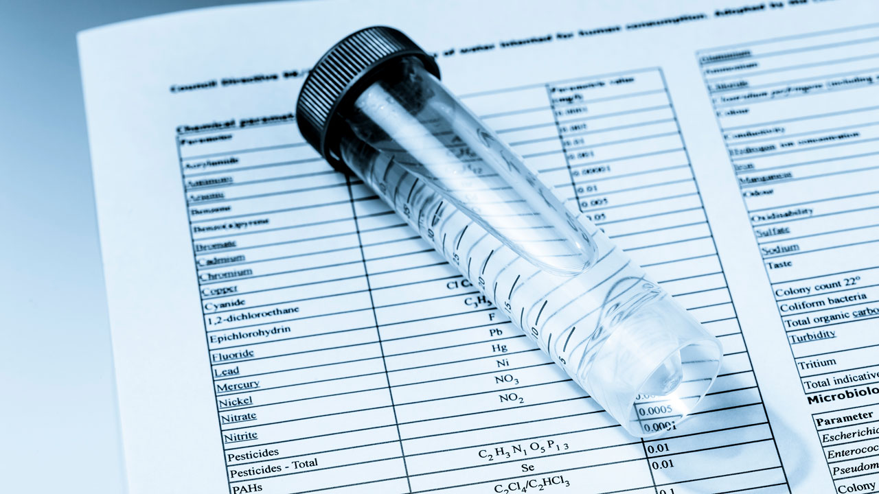 A vial of water on top of a paper with a grid showing several chemical contaminants in the form of a water quality report.