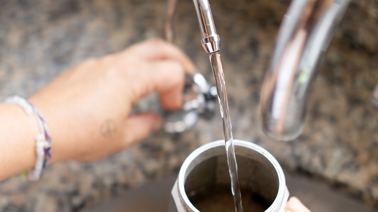 Hand turning on dedicated RO faucet to add water to a stainless steel coffee stovetop coffee maker.
