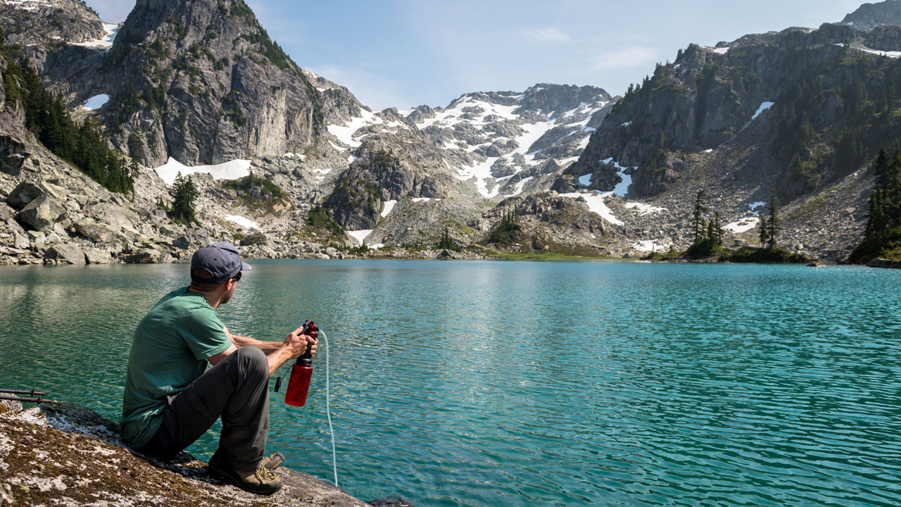 A person sitting by the side of a blue, high-elevation lake into a water bottle. surrounded by jagged peaks. He has a pump-style backcountry water filter and is filtering water from the lake