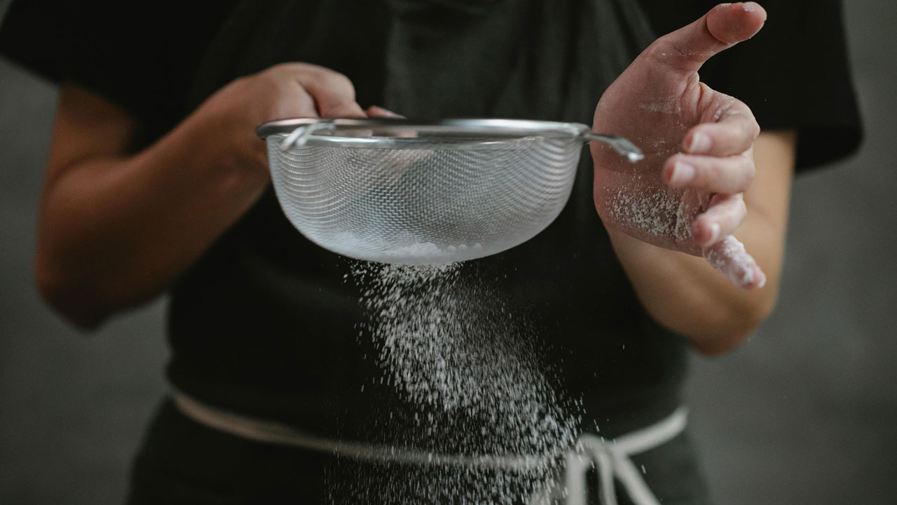Hands using a fine metal mesh sieve to sift flour in a kitchen demonstrating mechanical filtering