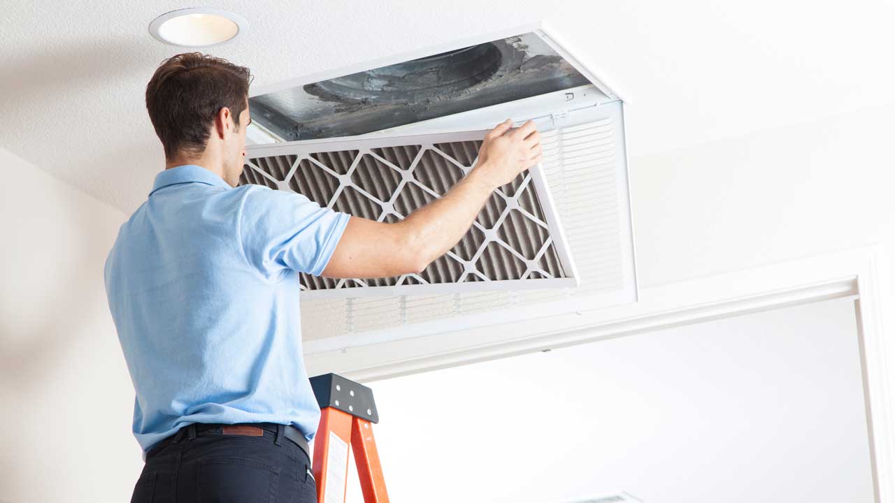 A male technician in a light blue shirt up on a ladder changing a filter in a ceiling mounted HVAC vent