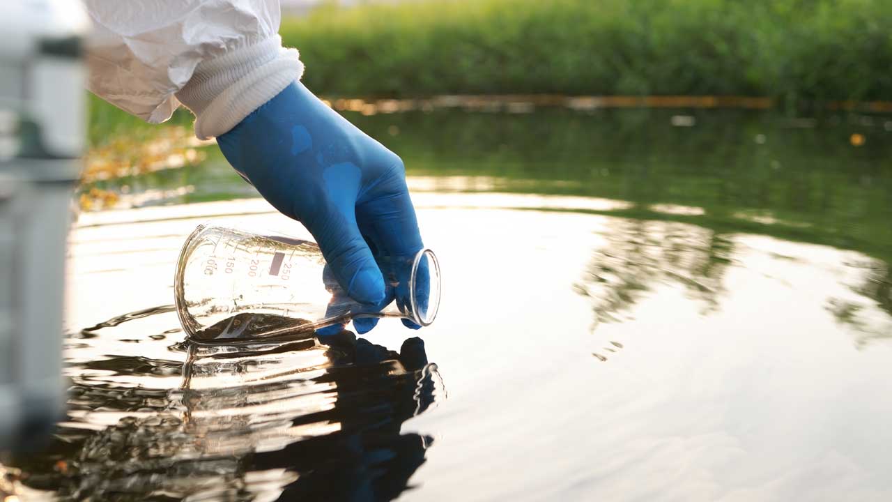 A hand in a rubber glove with a beaker dips into a water source for pollution testing