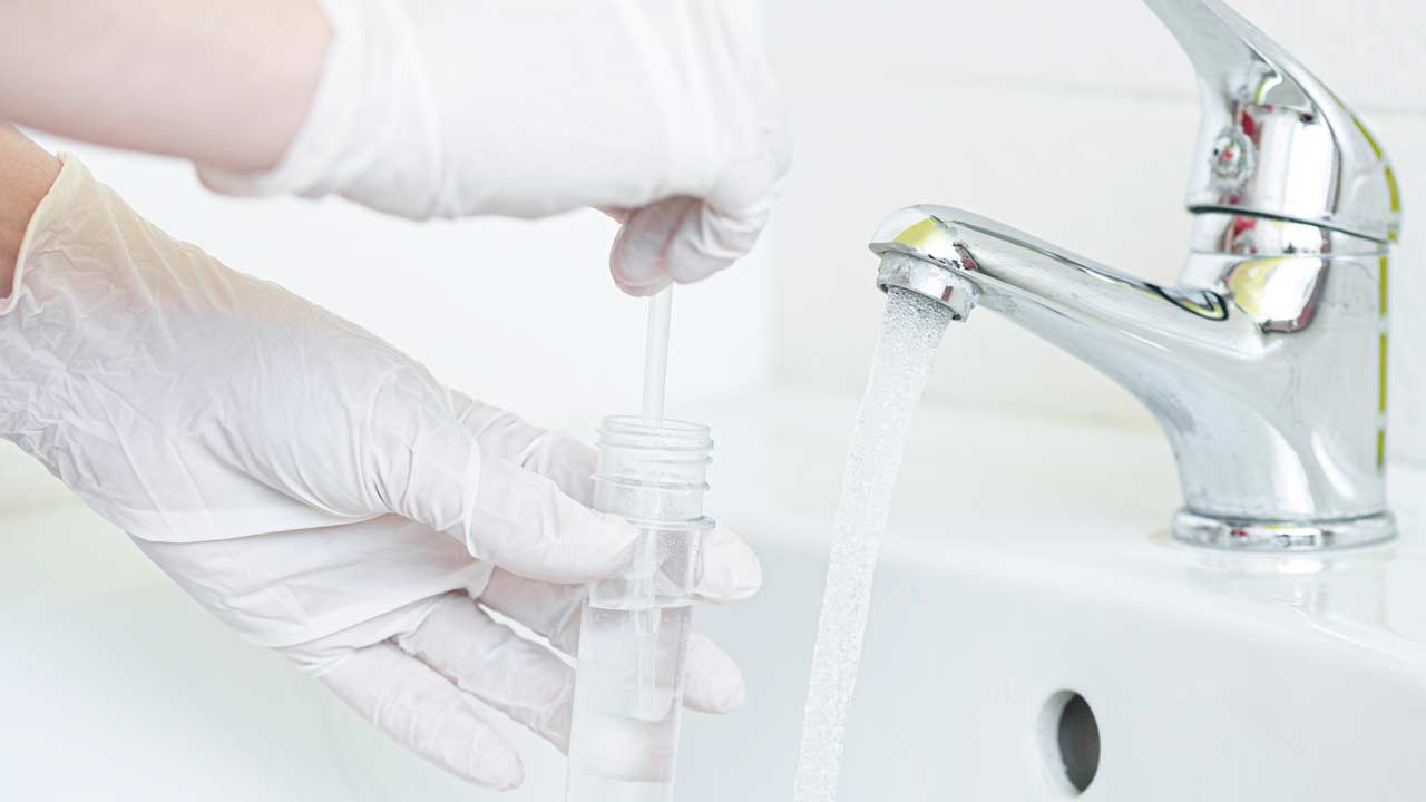 A gloved hand collects a water sample at a household faucet into a test tube