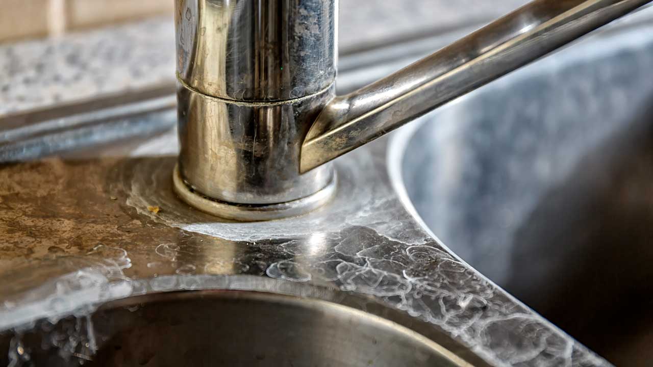 White mineral deposits called scale on a stainless steel kitchen faucet and sink