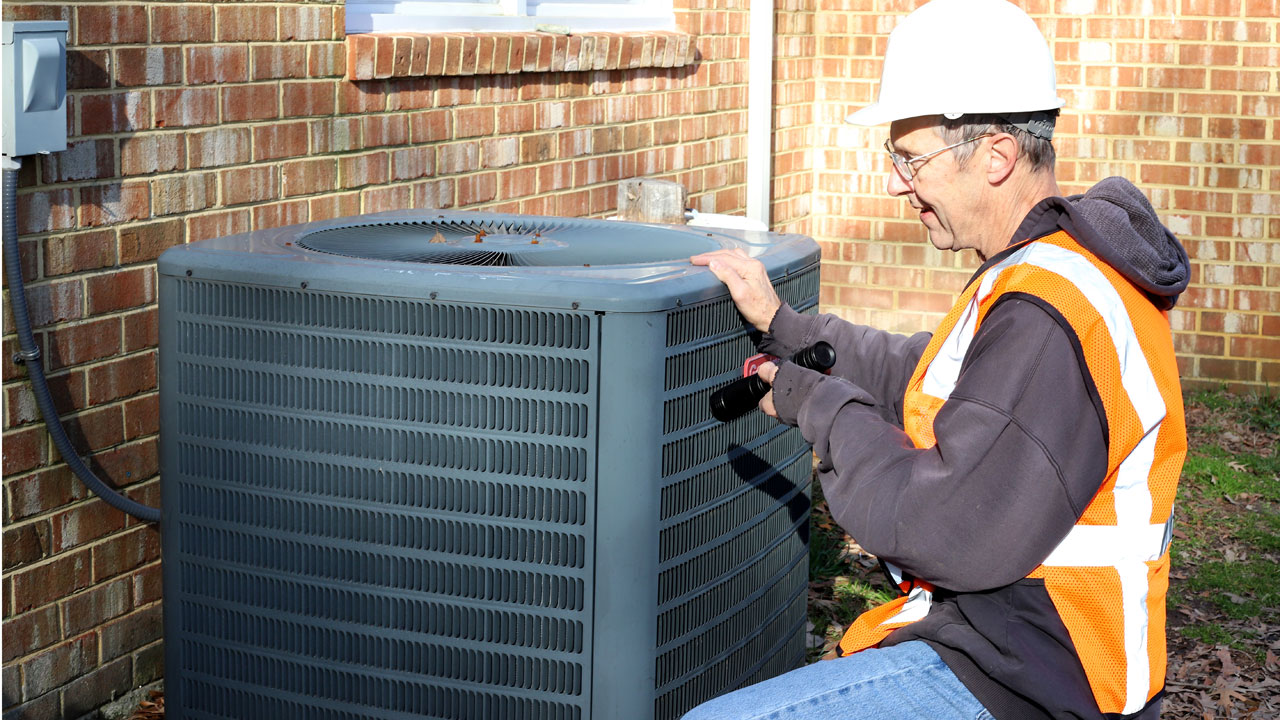A male HVAC technician with a reflective orange vest services the outdoor unit of an air conditioner HVAC system