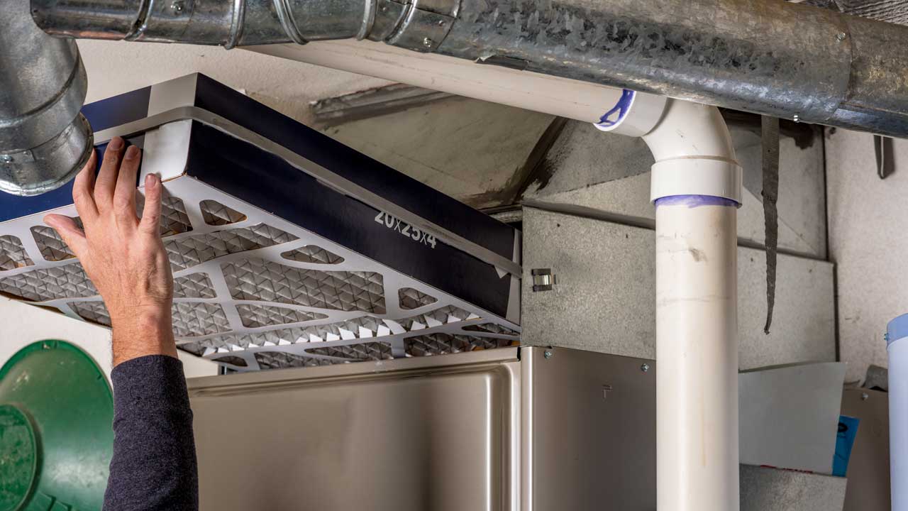 A hand reaches up high to install a clean 20" x 25" x 4" whole-house pleated air filter into a steel filter cabinet, like Argentine soccer player Diego Maradona's infamous Hand of God, except for a winning filter replacement 