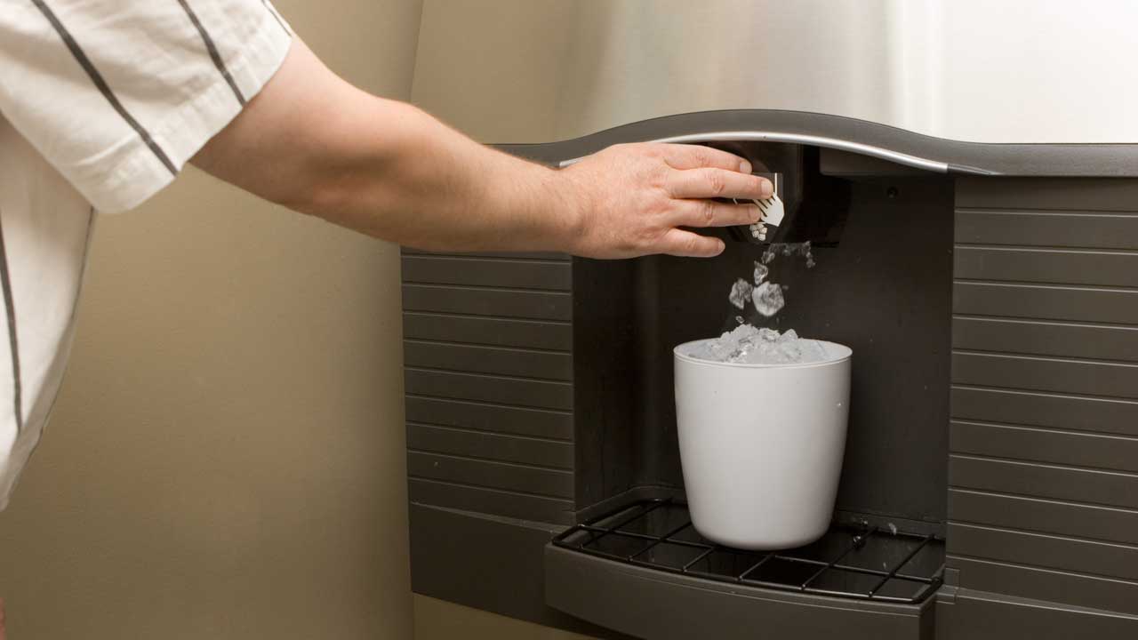 A hand presses the button on an ice machine to get cubed ice delivered to a white cup