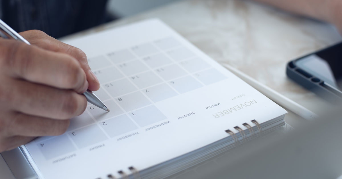 A hand marking a date on a printed desk calendar
