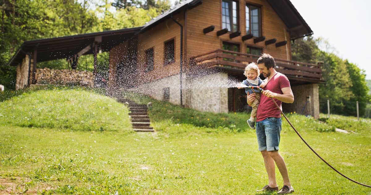 A man holding a toddler in one arm and a hose in the other, outside watering